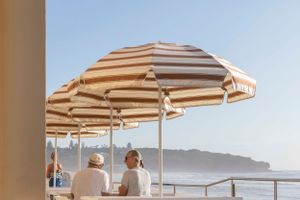 Striped beach umbrella shading beachgoers with coastal cliffs in the background.