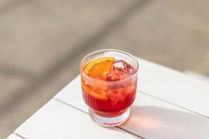 A bright red cocktail with ice in a lowball glass sits on a white surface casting long shadows.