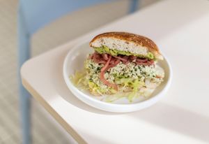 A gourmet sandwich filled with shaved meat, shredded lettuce and condiments on a fresh bun sits on a white plate.