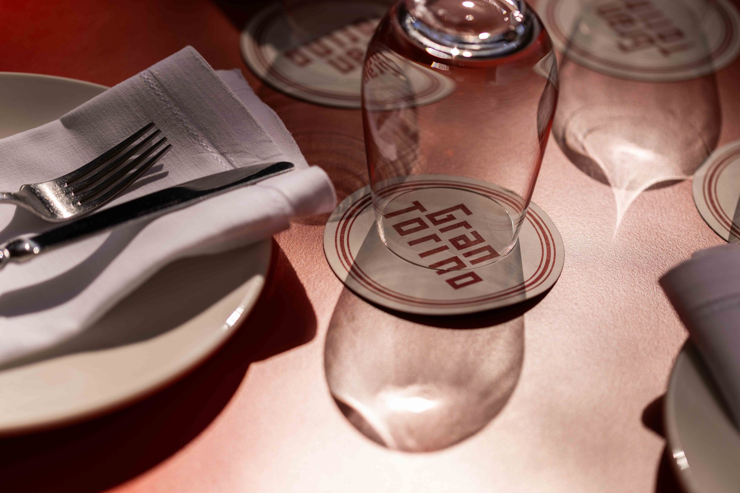 Gran Torino restaurant table setting with upturned wine glasses on branded coasters, white napkin, and fork on pink tablecloth.