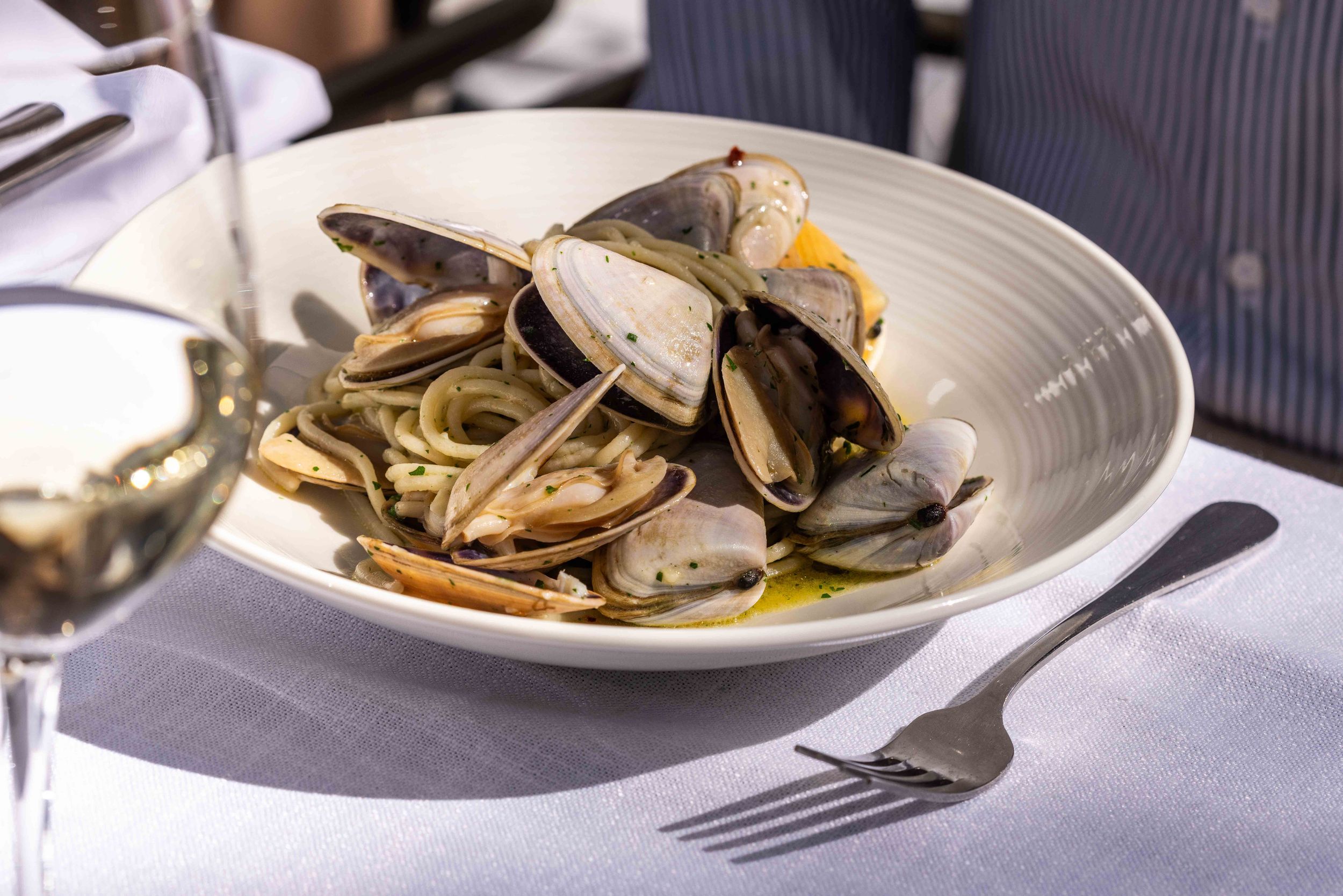 A white bowl of steamed mussels and clams with pasta in broth, served with white wine at a restaurant.