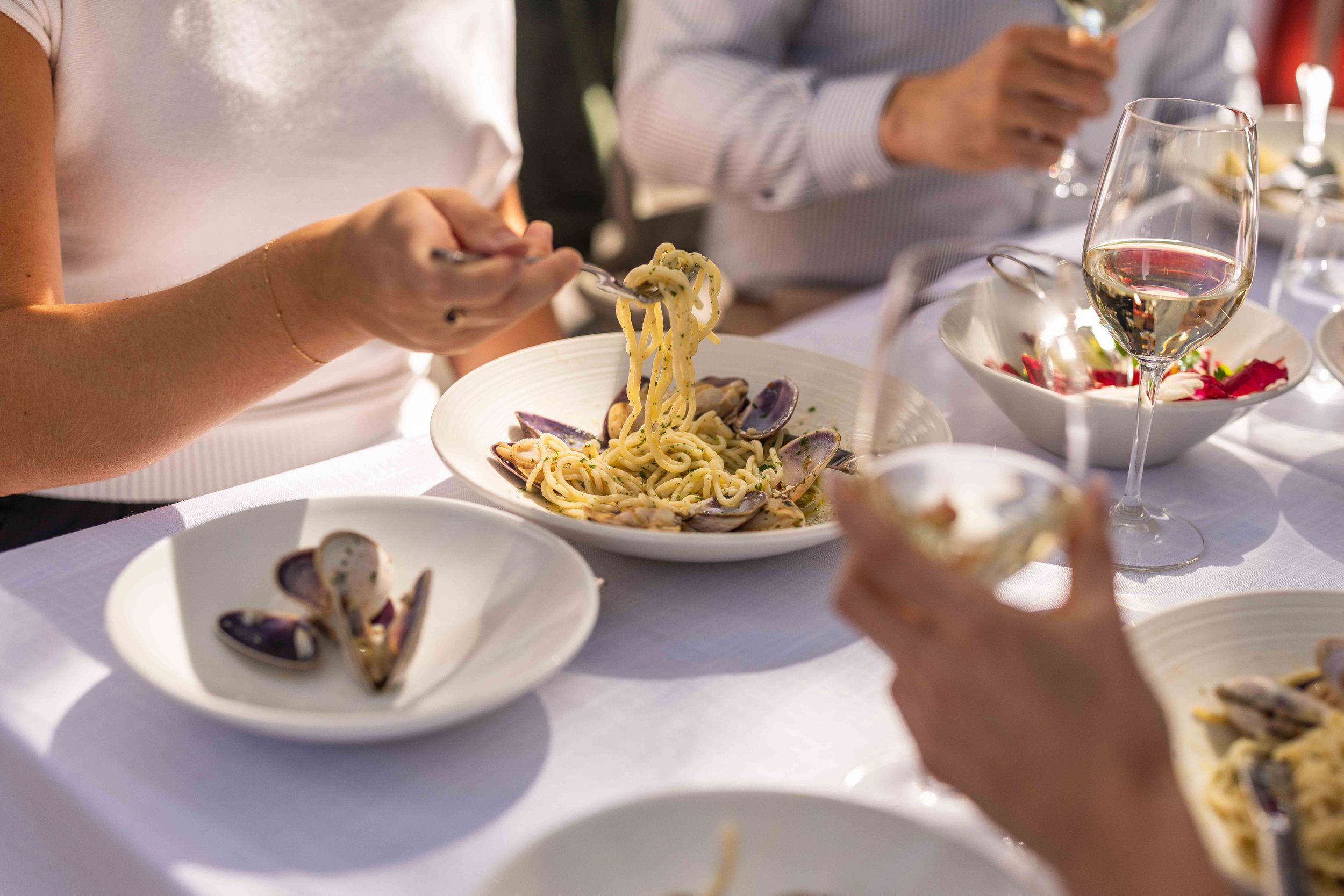 Elegant dining scene with pasta, mussels, and white wine glasses on a white tablecloth at a fine restaurant.
