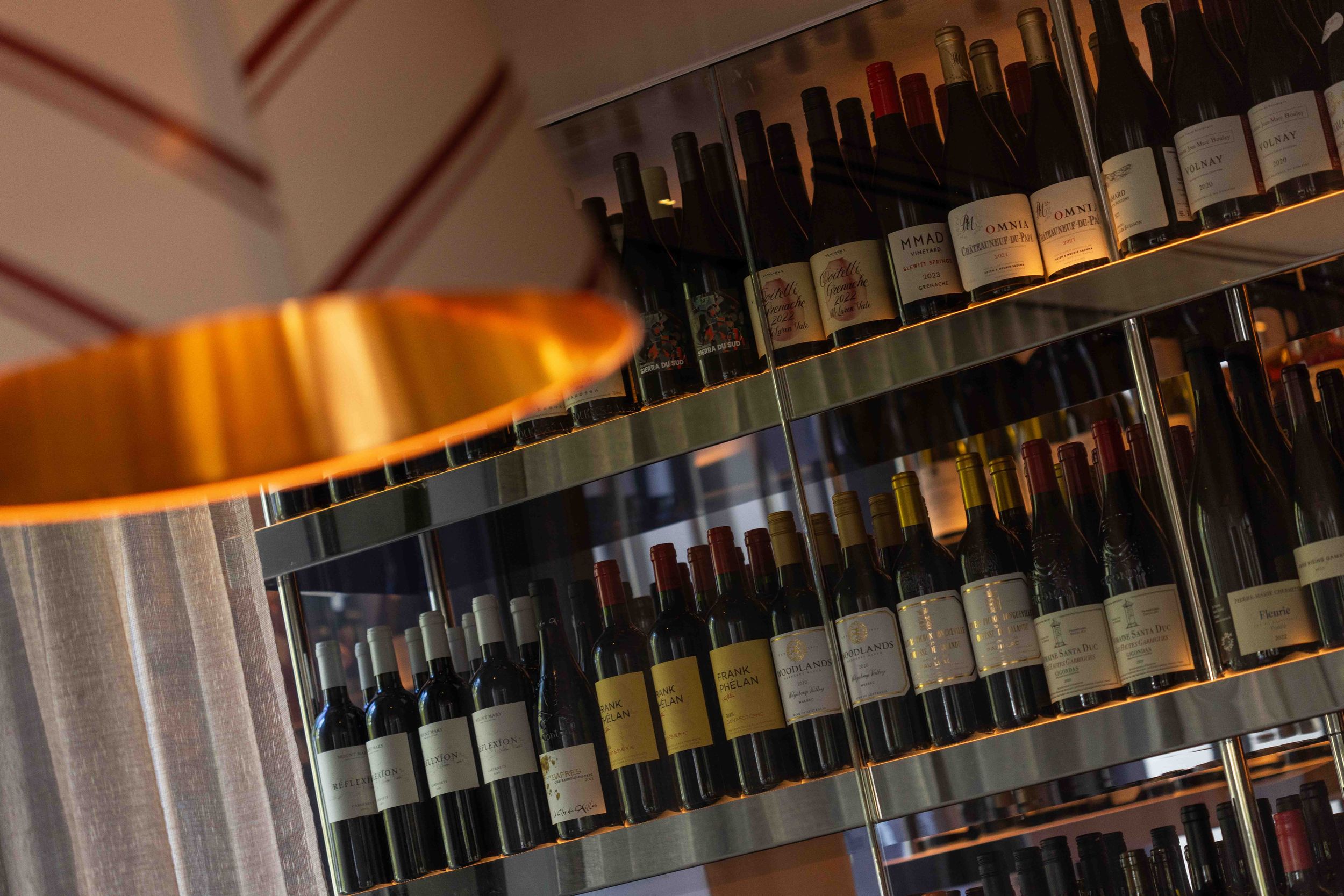 Copper pendant lamp beside shelves of wine bottles in a well-stocked wine bar or restaurant cellar.