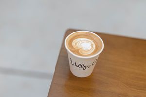 A paper coffee cup with latte art foam design sits on a wooden surface at Dialogue coffee shop.