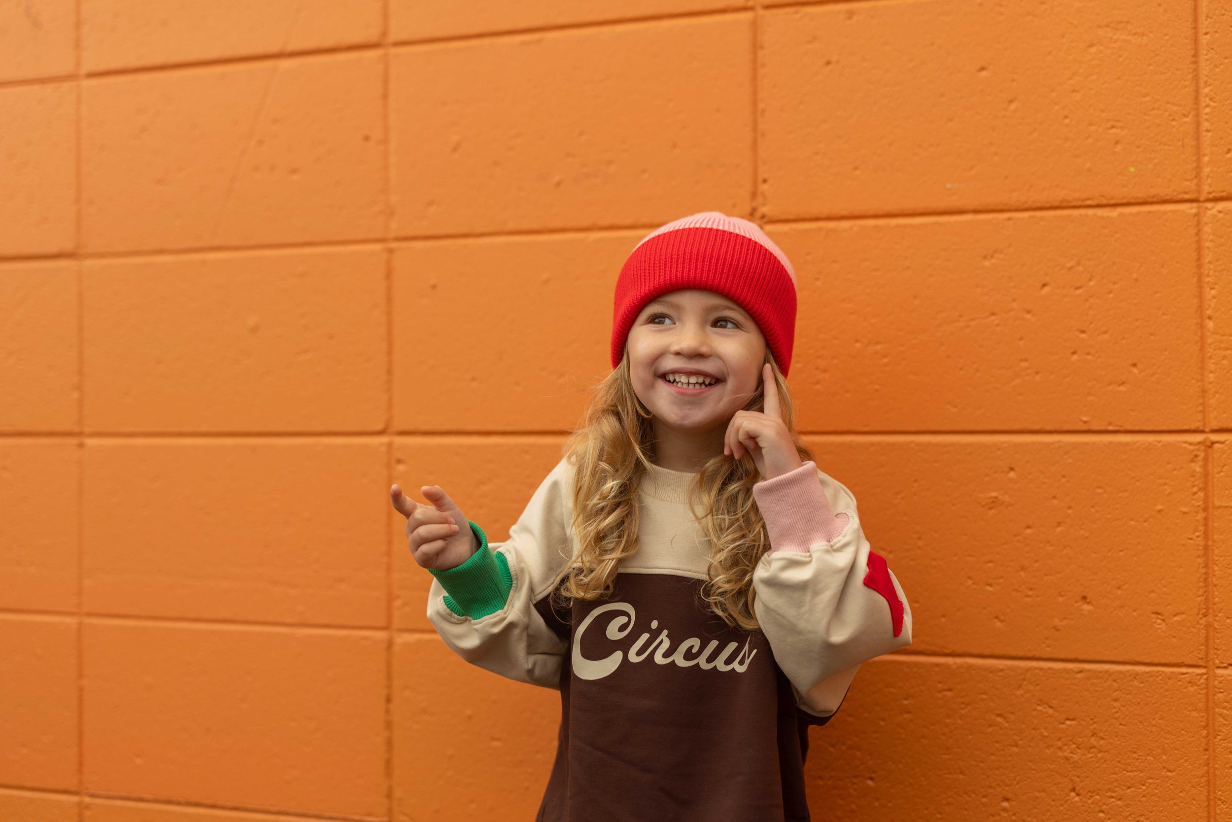 A smiling girl in a red beanie and Circus sweatshirt poses against a bright orange brick wall.