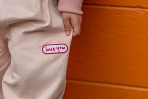 Close-up of pink sweatpants with a "love you" patch, held by a hand against an orange brick wall.