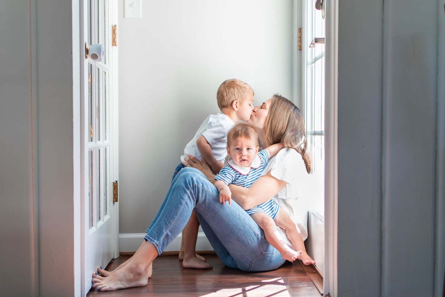 A mom and her two sons share a tender moment snuggled on the floor in front of the back door with light pouring in.