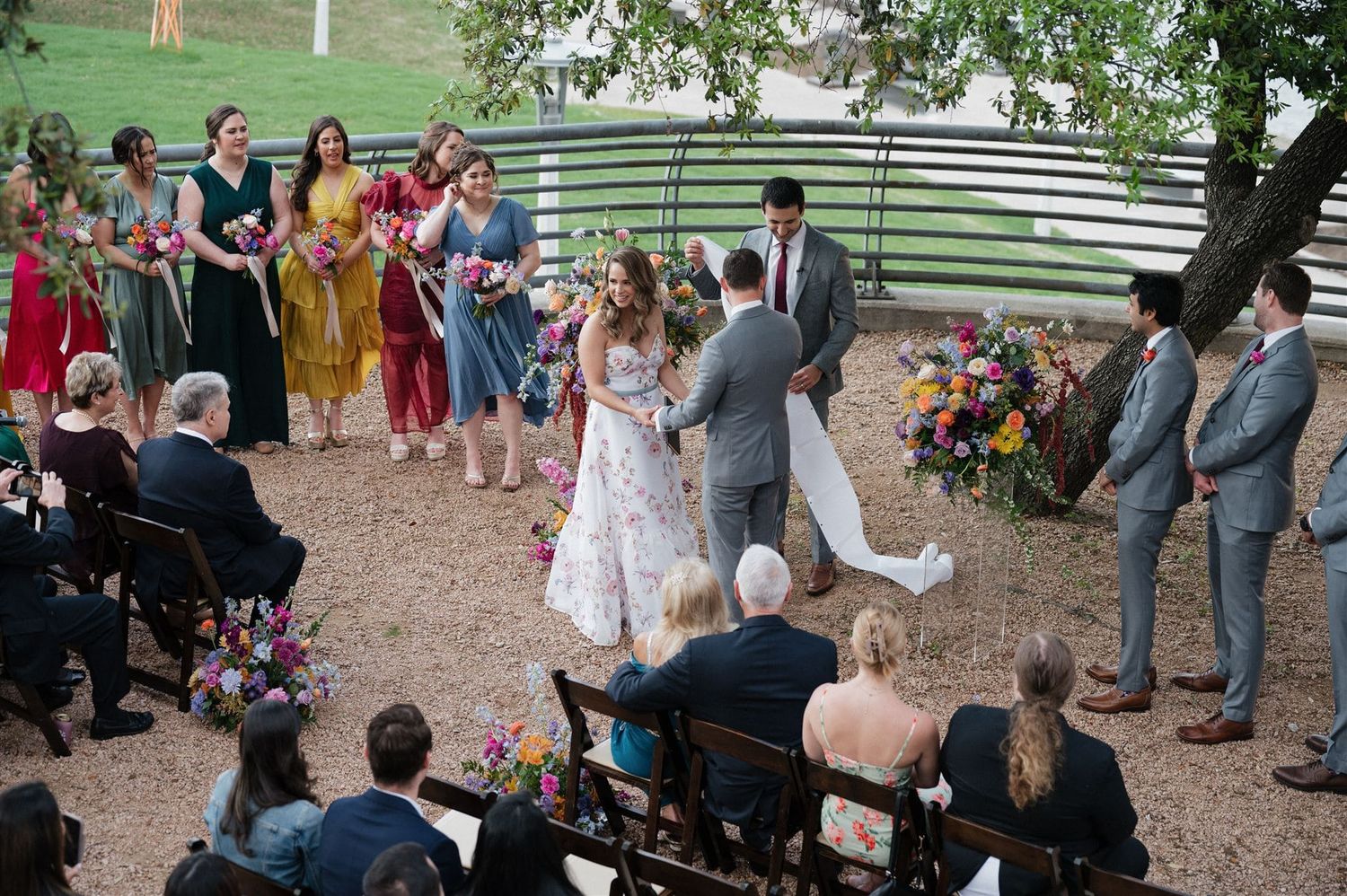 Outdoor wedding ceremony in progress with colorfully dressed guests and floral decorations.