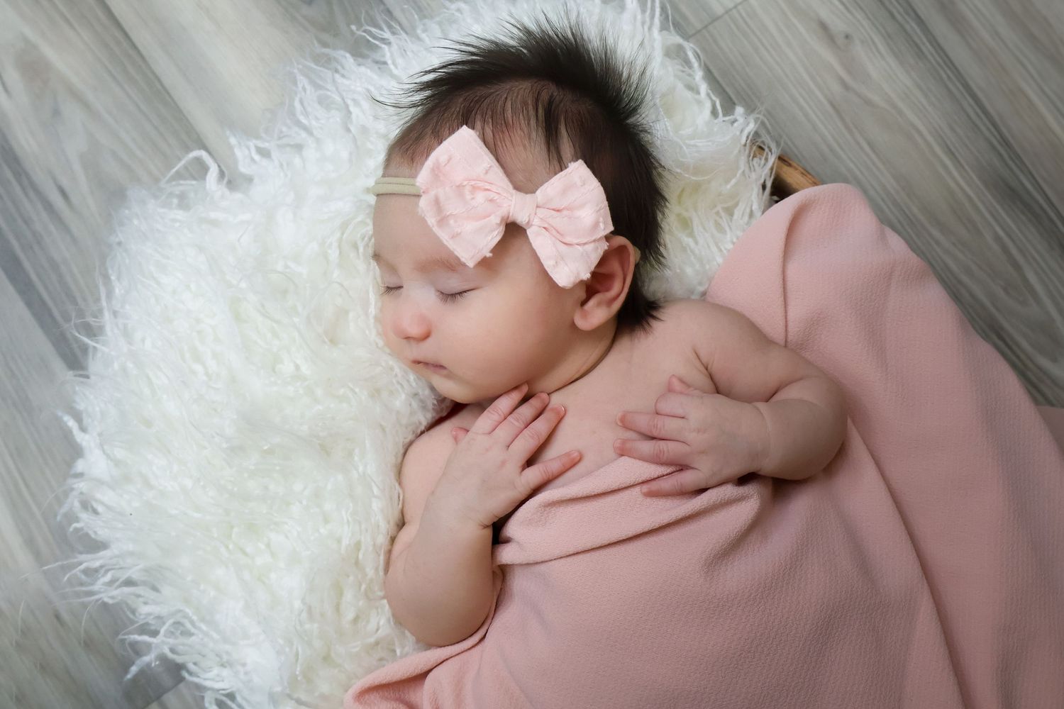 Sleeping newborn baby girl with a pink bow laying on a white fur with a pink blanket