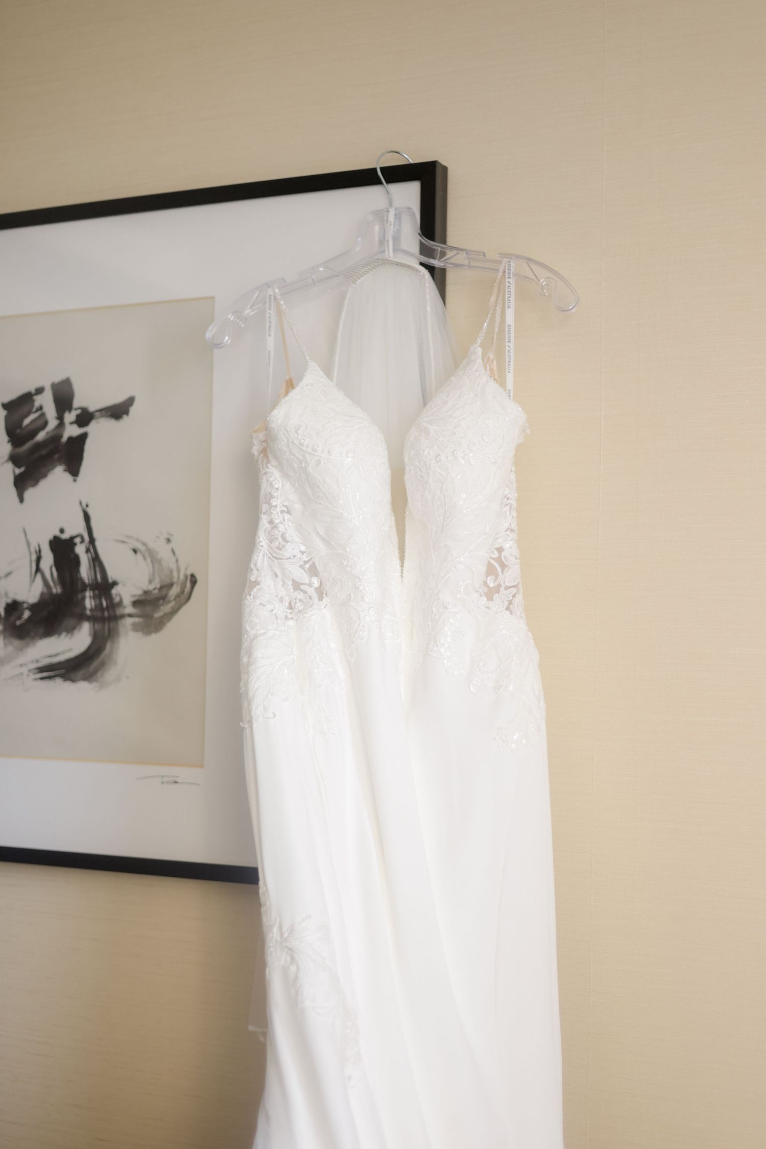 Elegant white wedding dress with lace details hangs against a beige wall near framed artwork.
