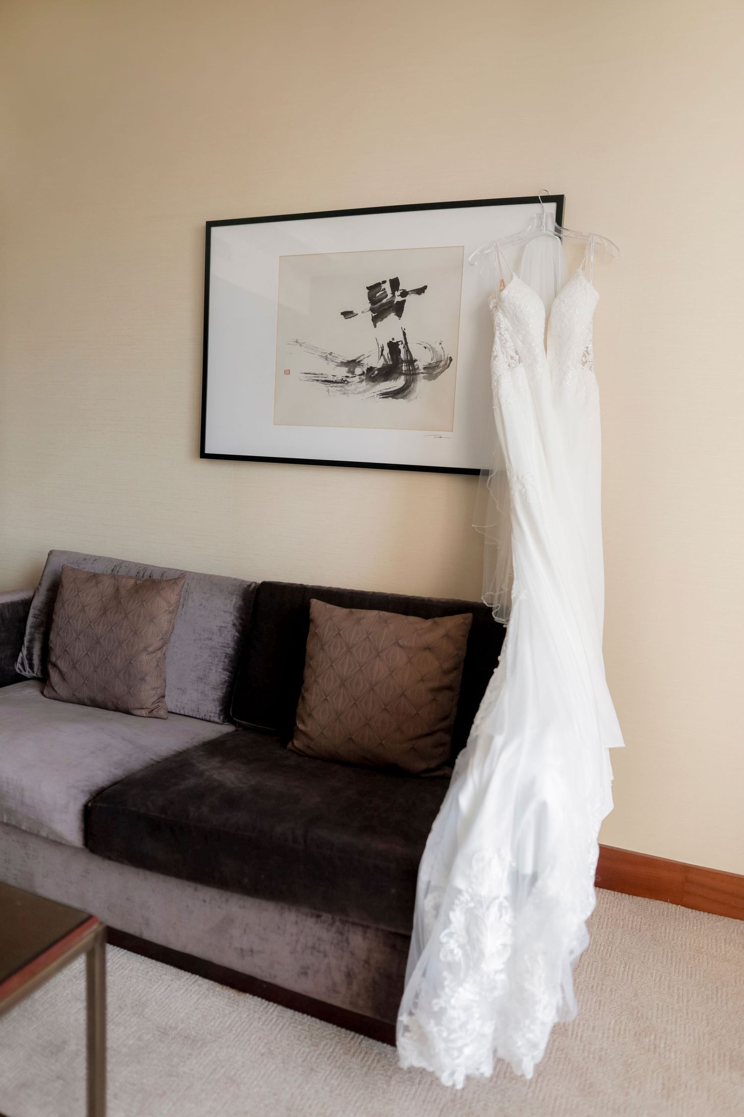 White lace wedding gown displayed against modern decor with gray sofa and framed artwork.