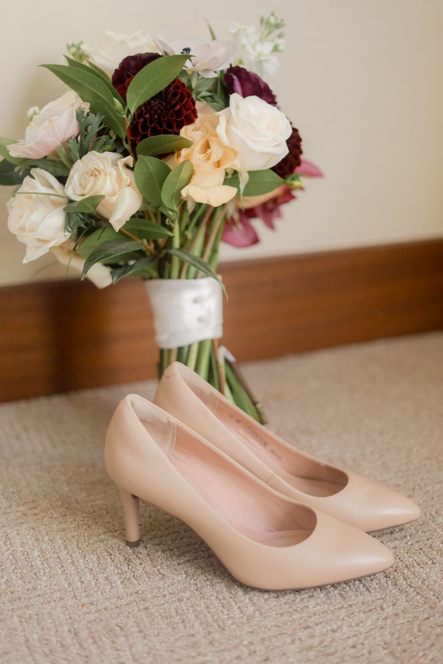 Bridal bouquet with burgundy and white flowers paired with nude heels on beige carpet.