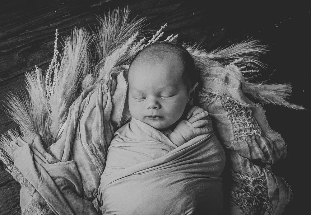 Black and white image showing a sleeping newborn wrapped in soft white fabric with wispy hair visible.