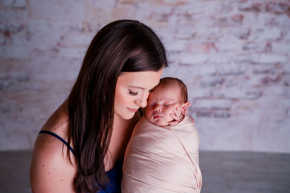 A tender newborn photography session shows parent and baby in soft lighting against a rustic brick wall background.