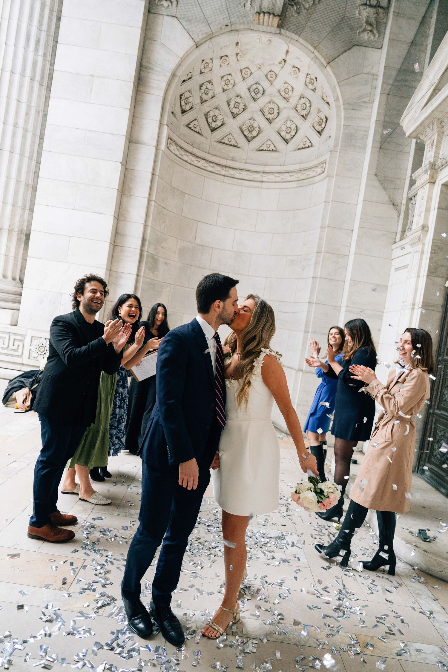 Couple shares romantic moment at courthouse wedding ceremony while friends celebrate with confetti in marble hallway.