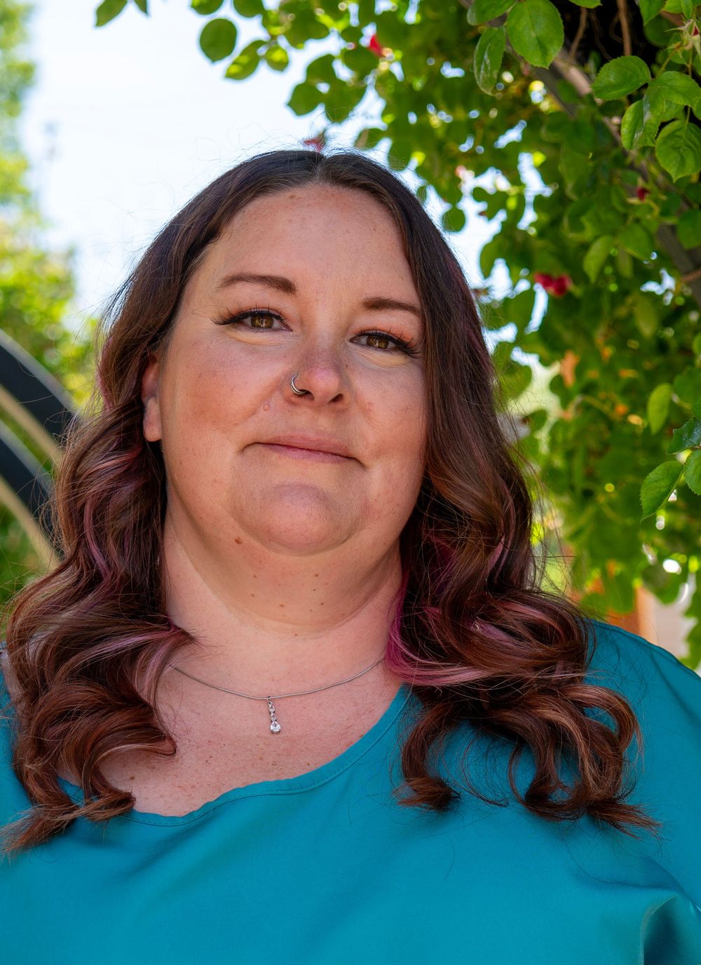 A woman with dark auburn wavy hair wearing a teal top and delicate necklace poses outdoors near green foliage.