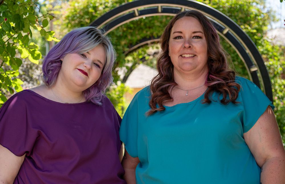 Two women smiling outdoors near a garden arch, one in purple top with lavender hair, one in teal top.