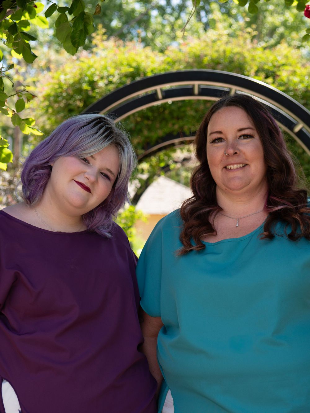 Two women smiling outdoors, one with purple hair in purple dress, other in teal top, posed under garden arch.