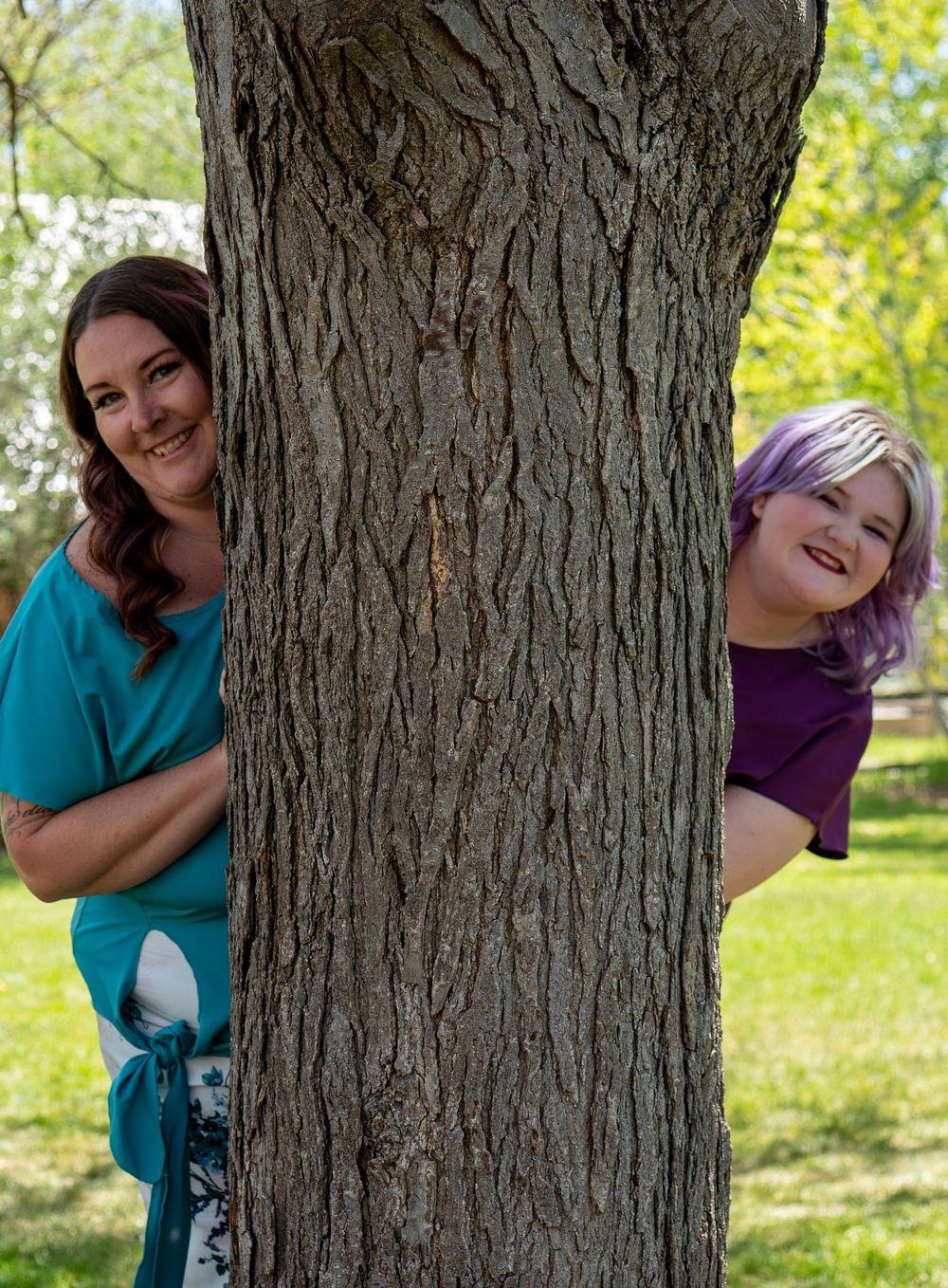 Two women peeking playfully from behind a large tree trunk in a sunny green park, one in teal and one in purple.