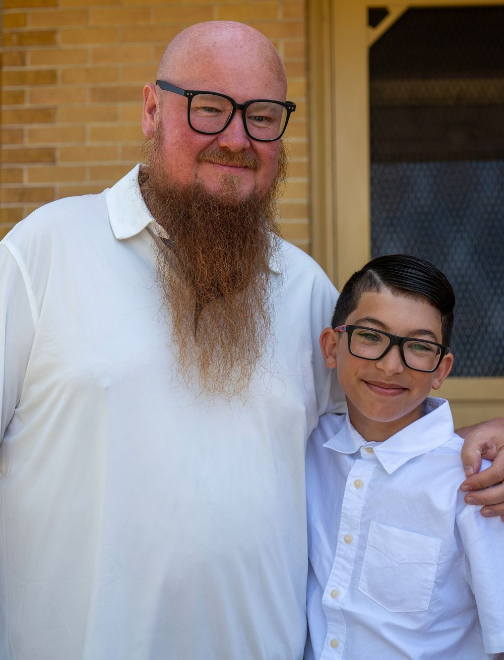 Tall bearded man with long red beard and glasses poses with young boy in white shirts outside brick building.