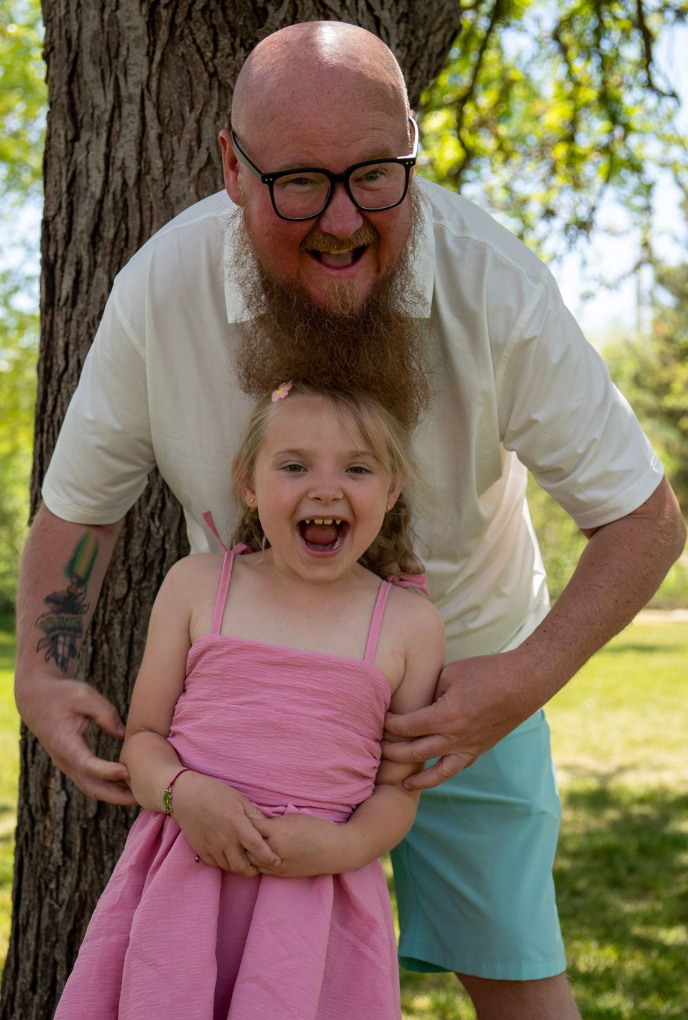 Bald bearded man with glasses laughing with young girl in pink dress outdoors by tree.