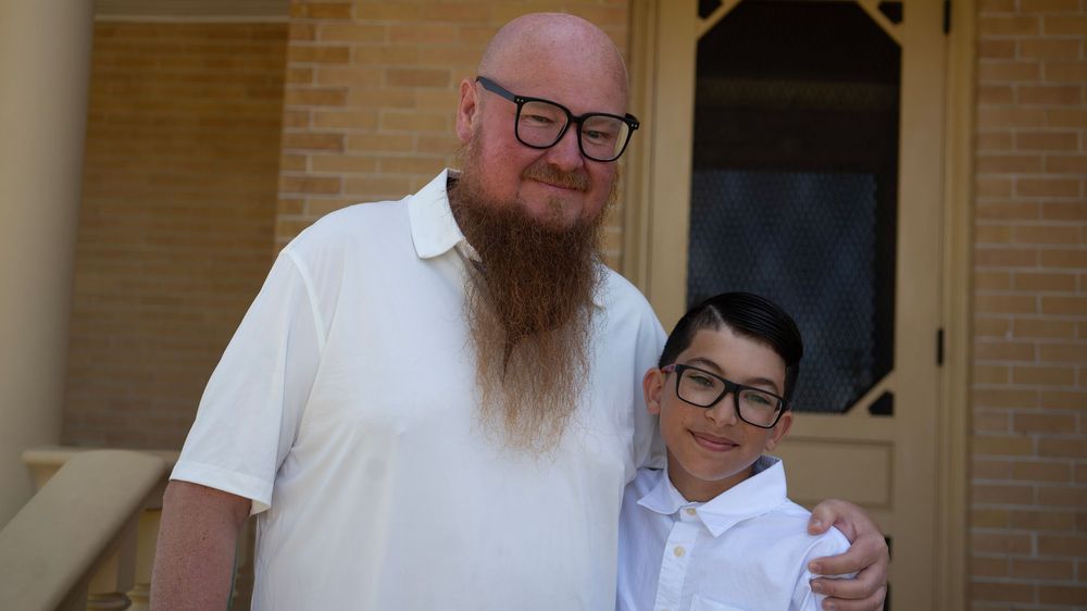 A large bearded man in a white polo poses with a young boy in a white shirt outside a brick building.