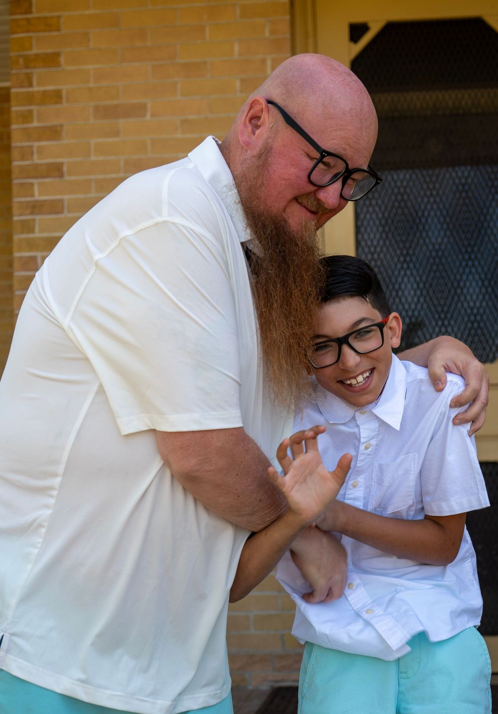 Bald bearded man in white polo hugging smiling boy with glasses outdoors near brick building.
