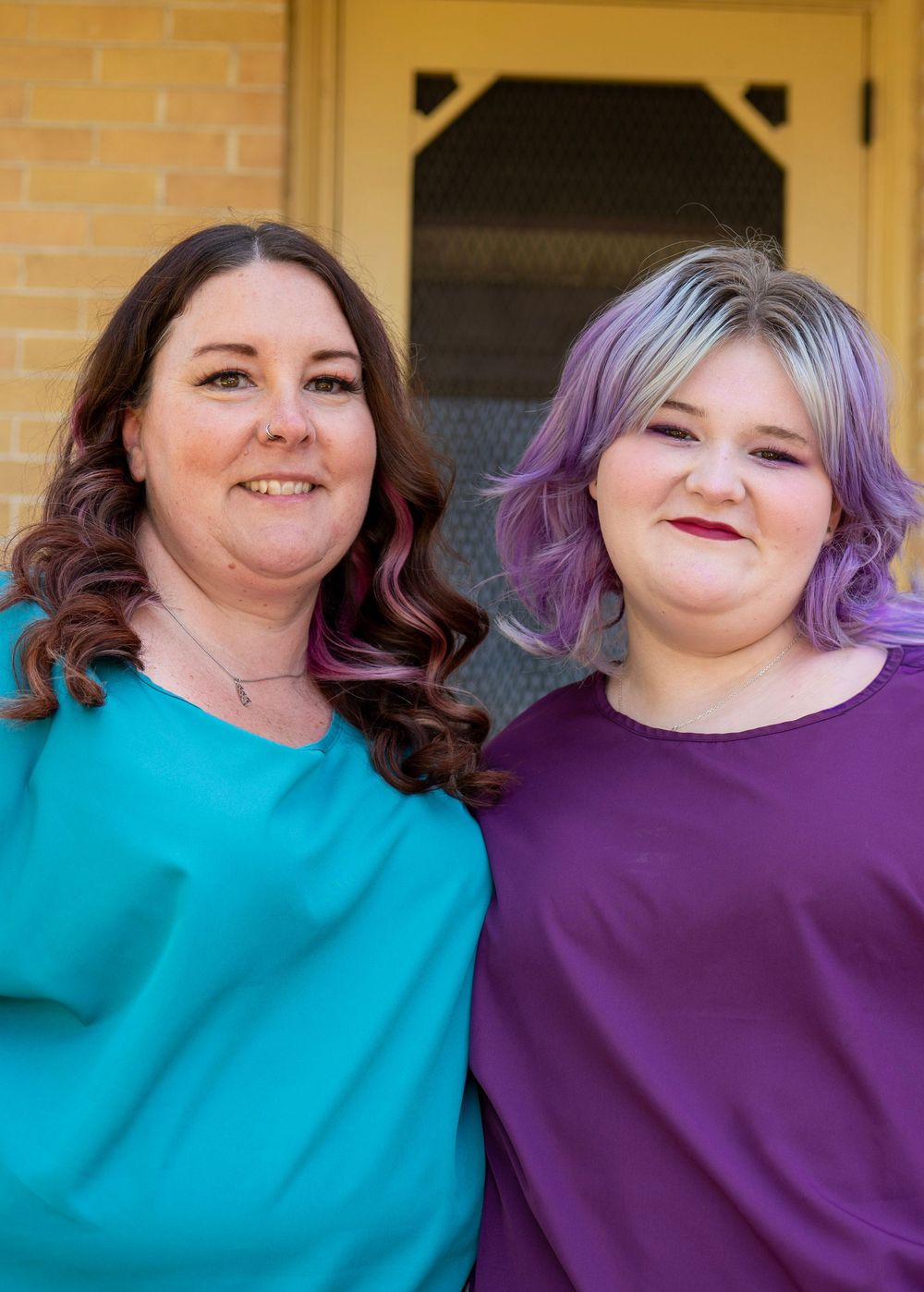 Two women with purple-tinted hair smiling together, one in teal and one in purple top, standing outside a brick building.