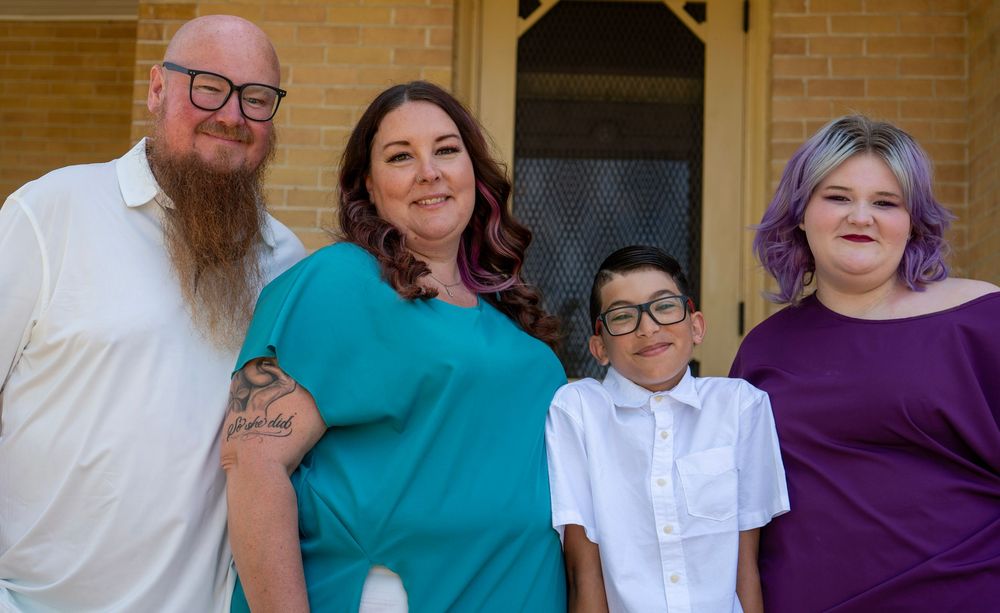 Family of four smiling together outside brick home, man with beard, woman in teal, boy in white, girl in purple.