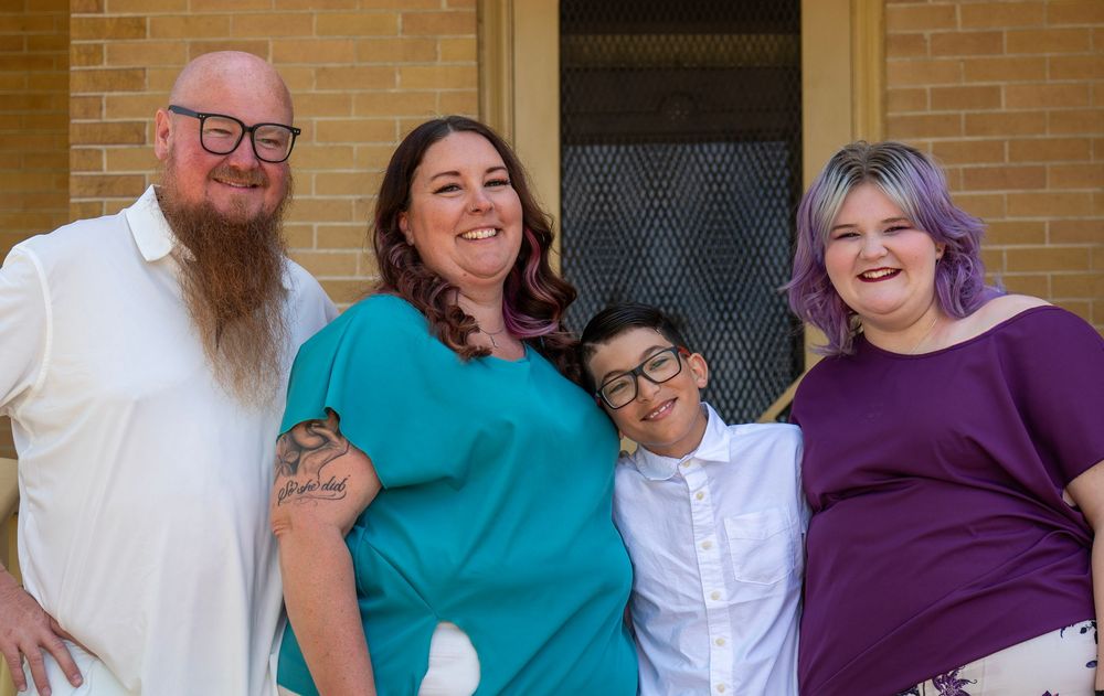 A joyful family of four poses outdoors near a brick building, smiling together in colorful casual attire.