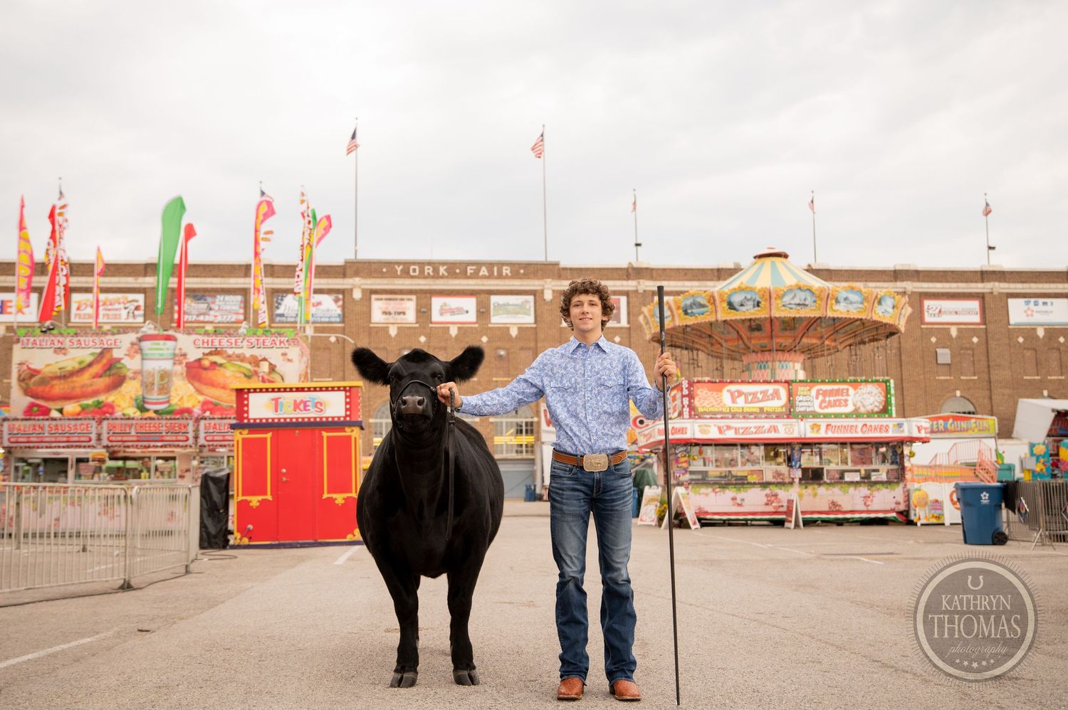 A person stands with a large black show cow at a fairground with carnival booths and flags in background.