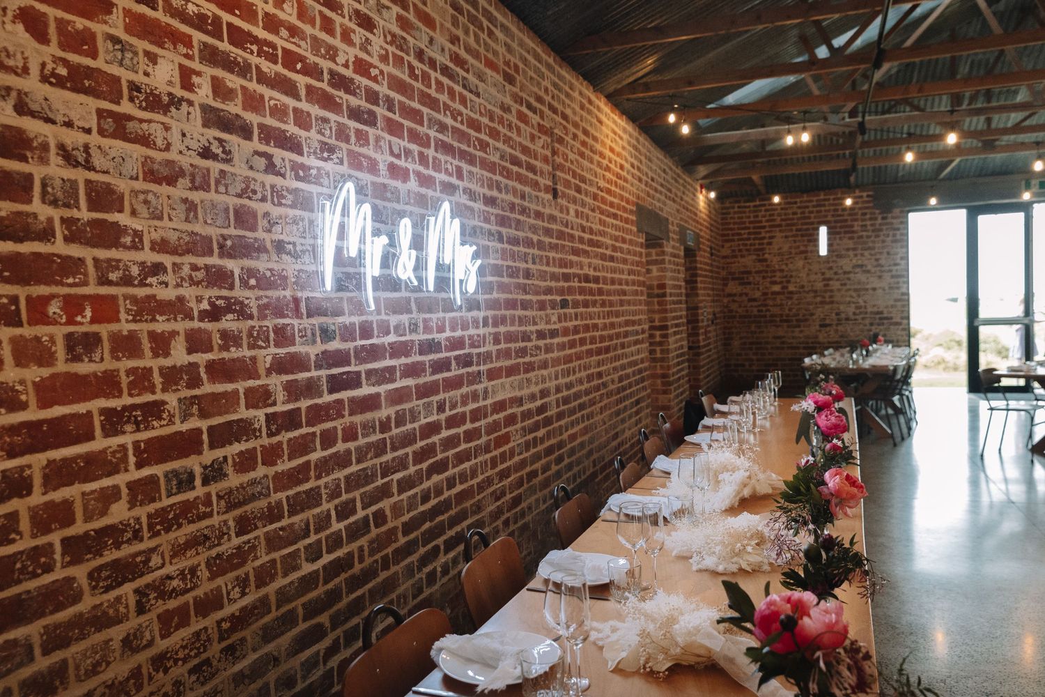 Long reception table set against brick wall with neon lighting and floral centerpieces.