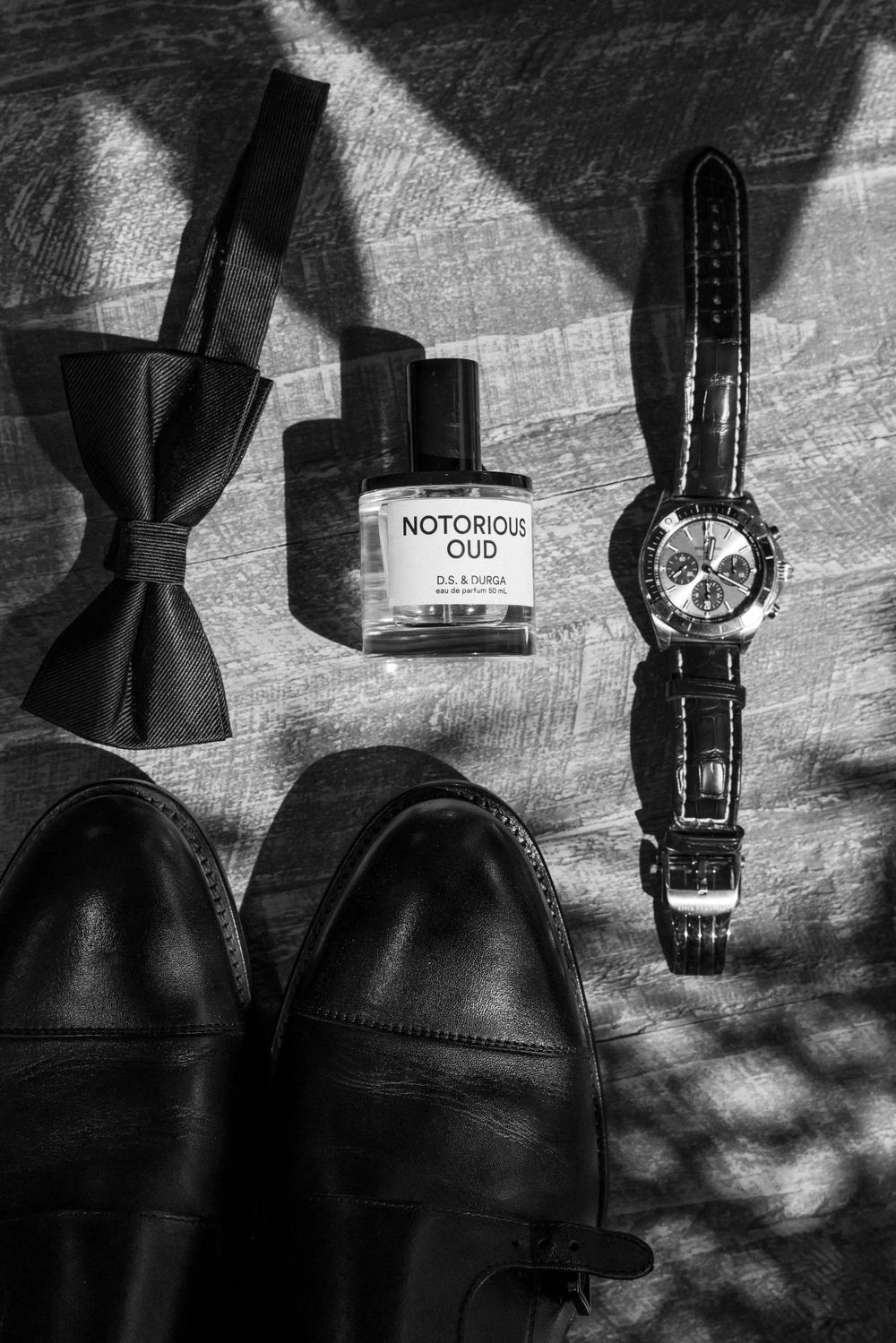 Groom's accessories including bow tie, dress shoes, cologne and watch arranged on dark surface.