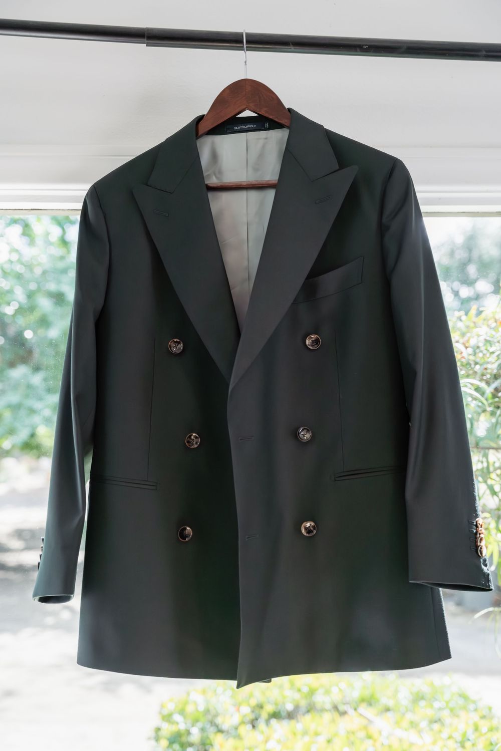 Black double-breasted formal suit jacket hanging on wooden hanger against bright window.