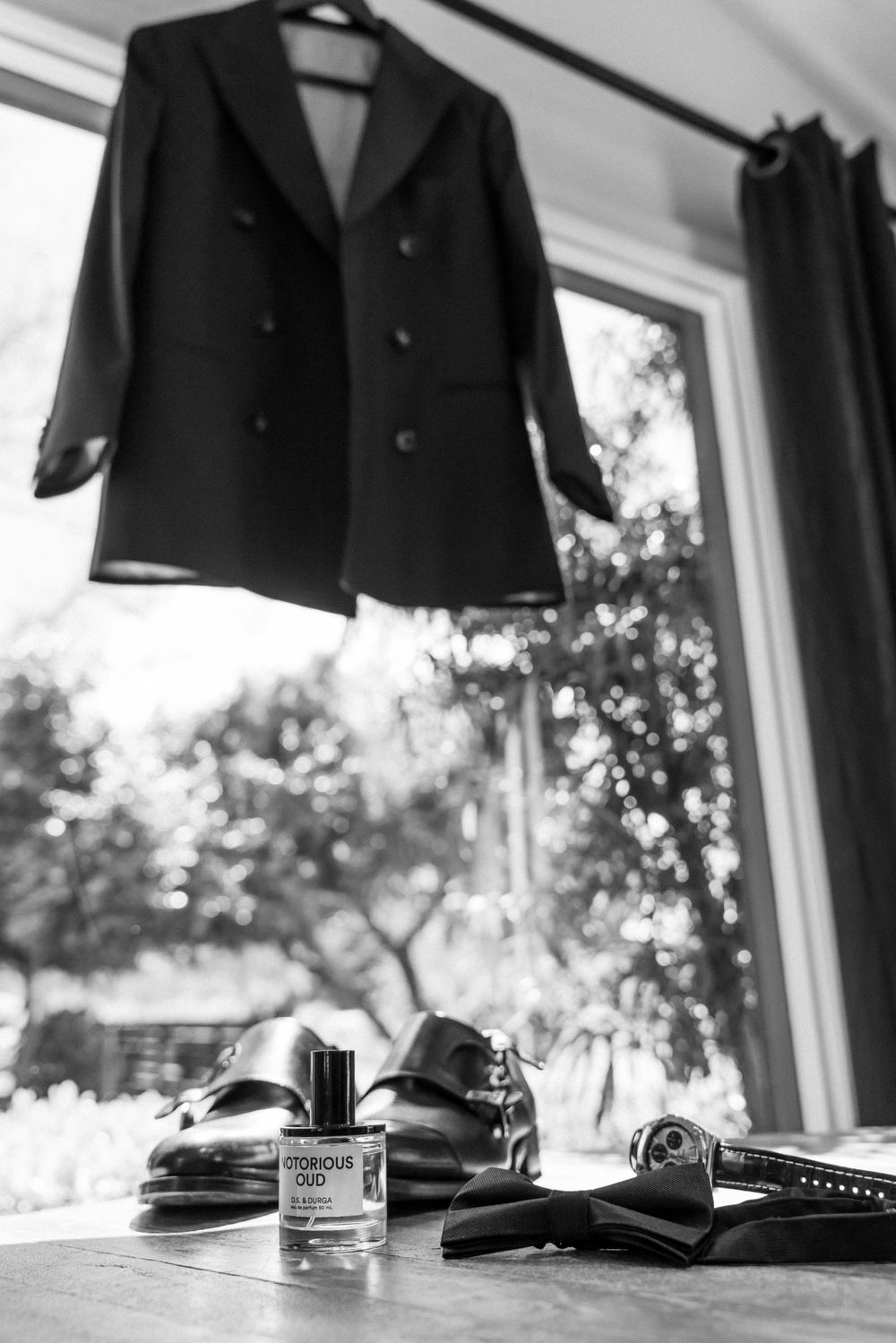 Formal black suit jacket with accessories and shoes arranged by window in black and white.