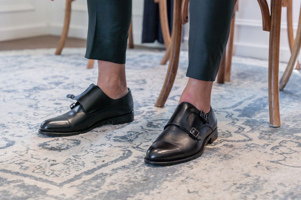 Black leather monk strap dress shoes worn with dark pants on patterned carpet.