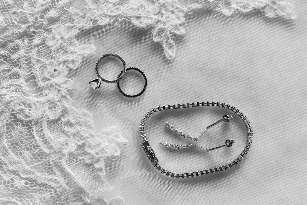 Wedding rings and lace accessories arranged artistically in black and white on delicate lace fabric.