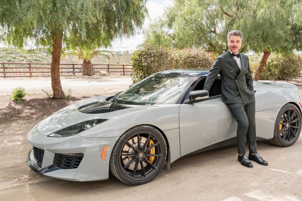 A sleek silver sports car parked outdoors with someone posing beside it in formal attire.