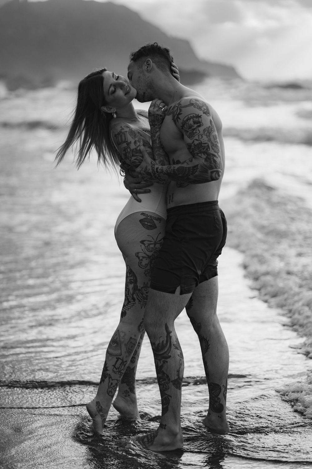 A romantic black and white beach photo of a tattooed couple embracing at the water's edge.