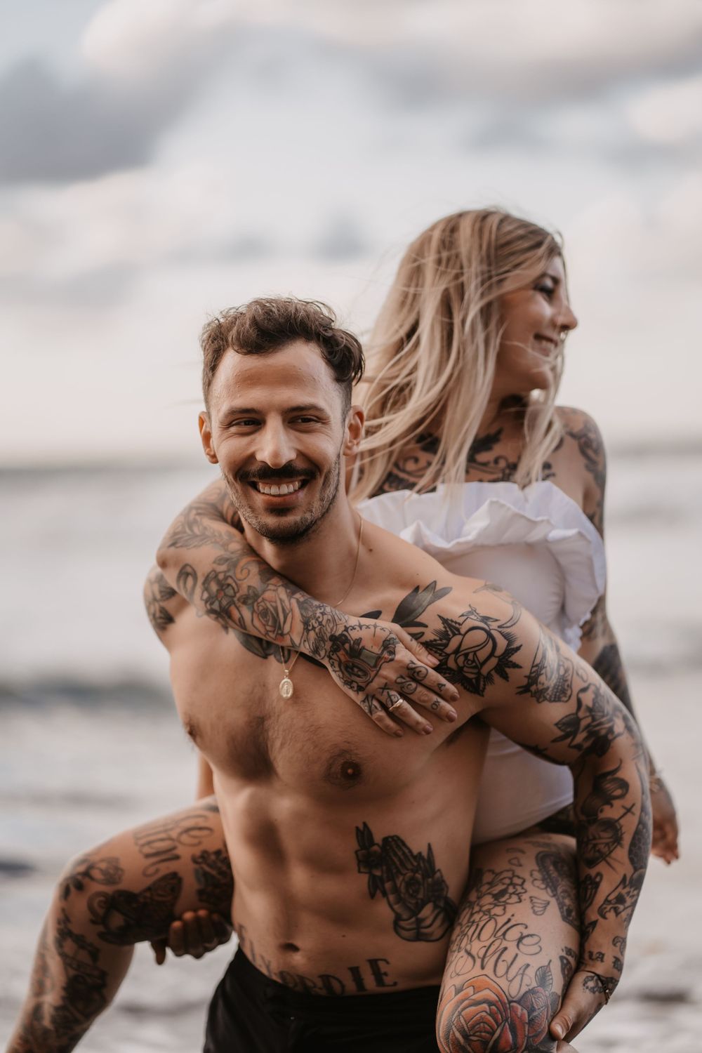 A couple embraces and laughs together on a beach during sunset, showcasing visible tattoos and casual attire.