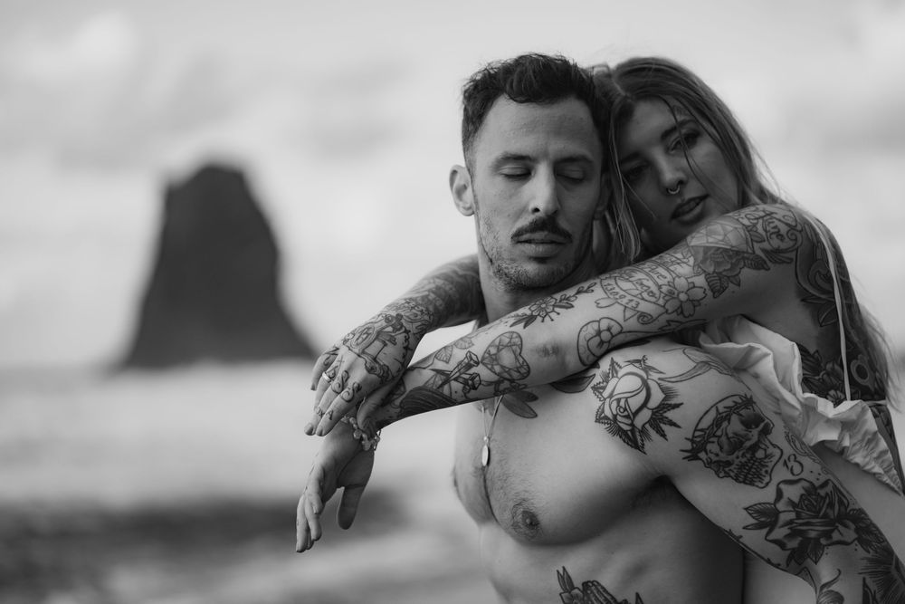 Black and white artistic photo of a tattooed couple embracing on a beach with a rock formation in the background.