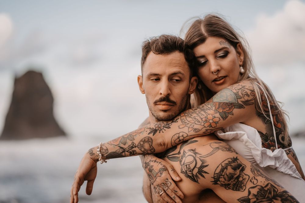 A couple embraces on a rocky beach while waves crash against the shore in the background.