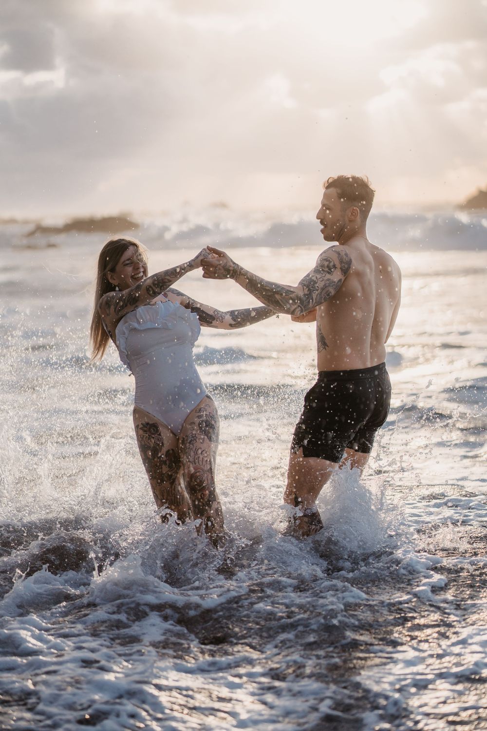 Two people playfully splashing and running through ocean waves at sunset while water sprays around them.