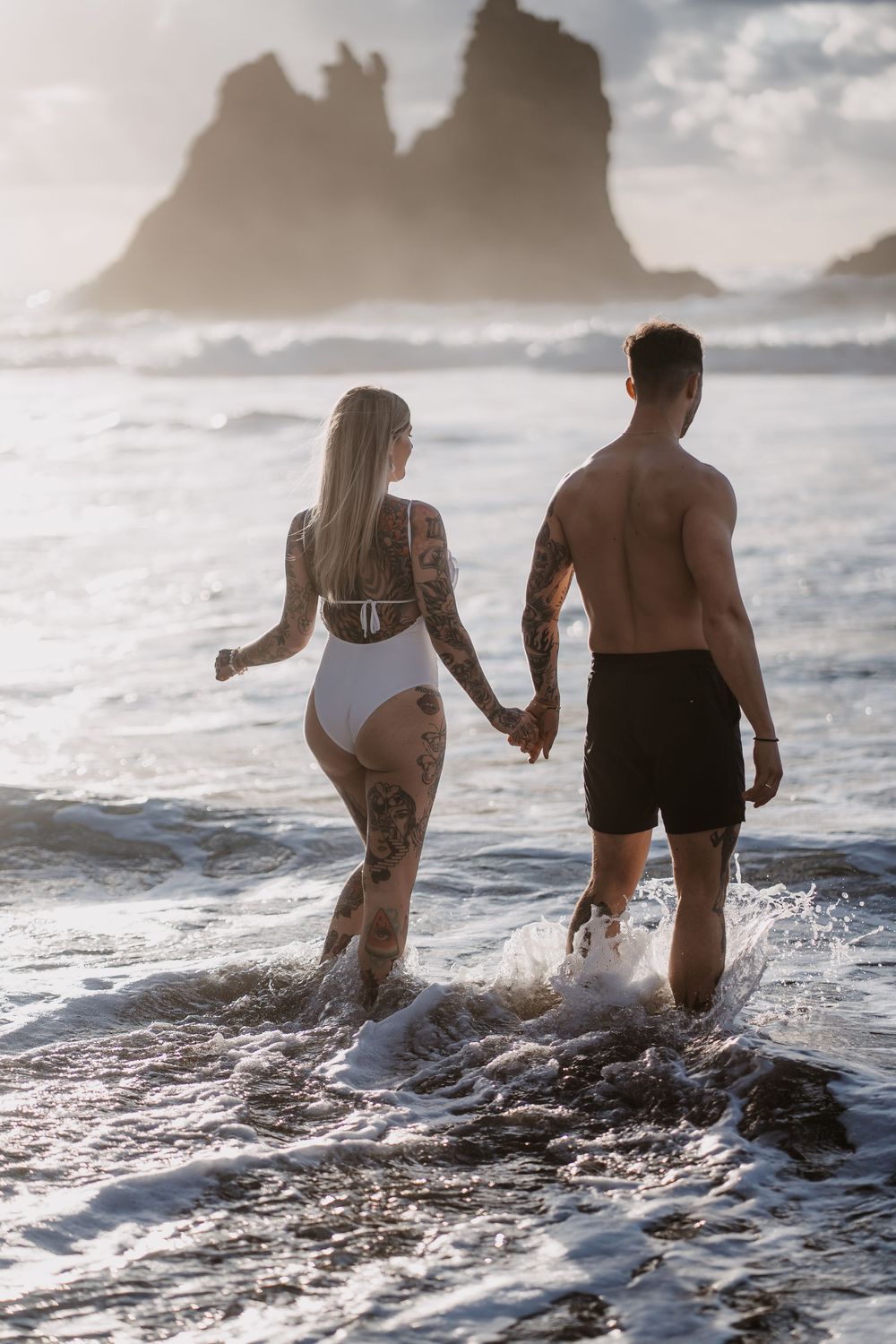Two people wade into the ocean waves at sunset, creating splashes as they venture deeper into the water.