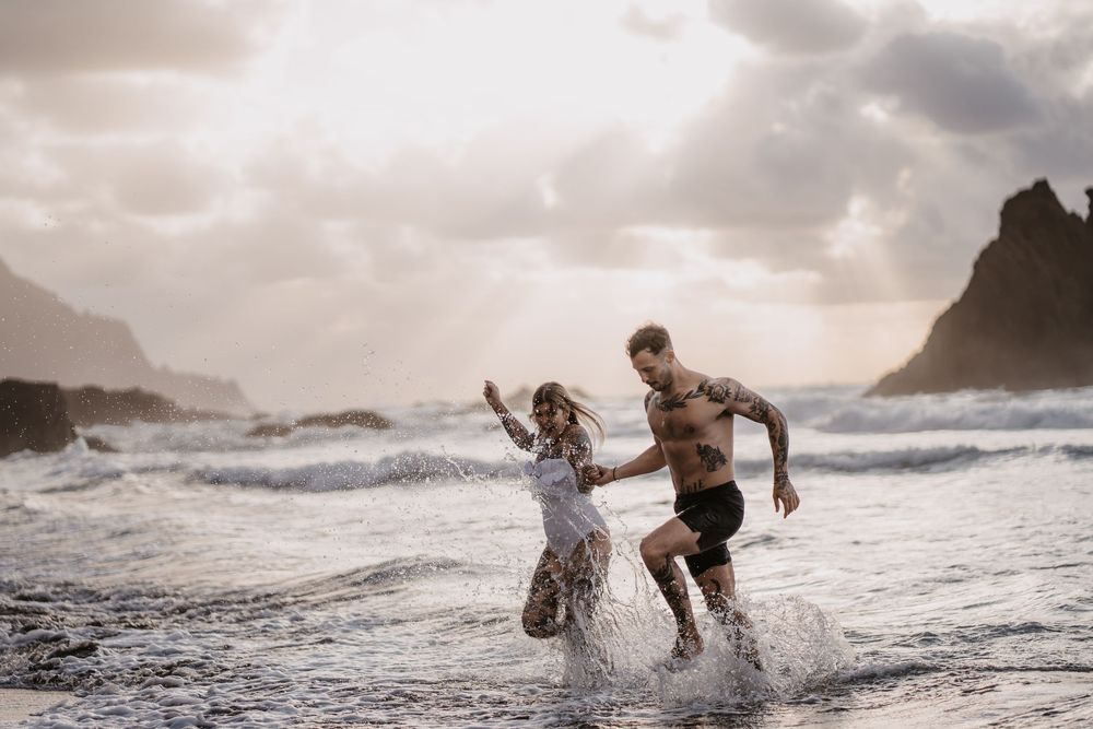 A sequence of silhouetted figures playing and splashing in dramatic ocean waves at sunset along a rocky coastline.