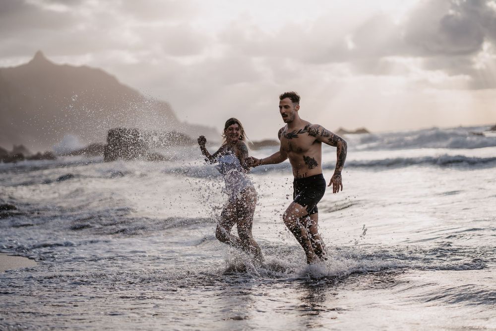 People splash and run through ocean waves at sunset along a dramatic coastline with mountains in the background.