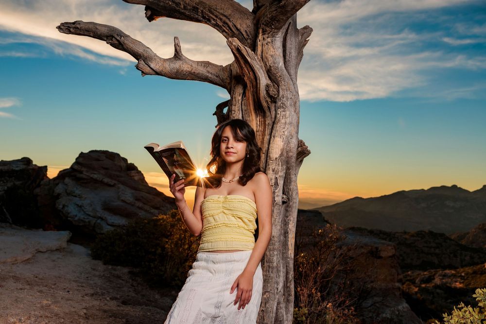 senior girl reading a book leaning on a tree on Mt. Lemmon with mountain views and a sun burst