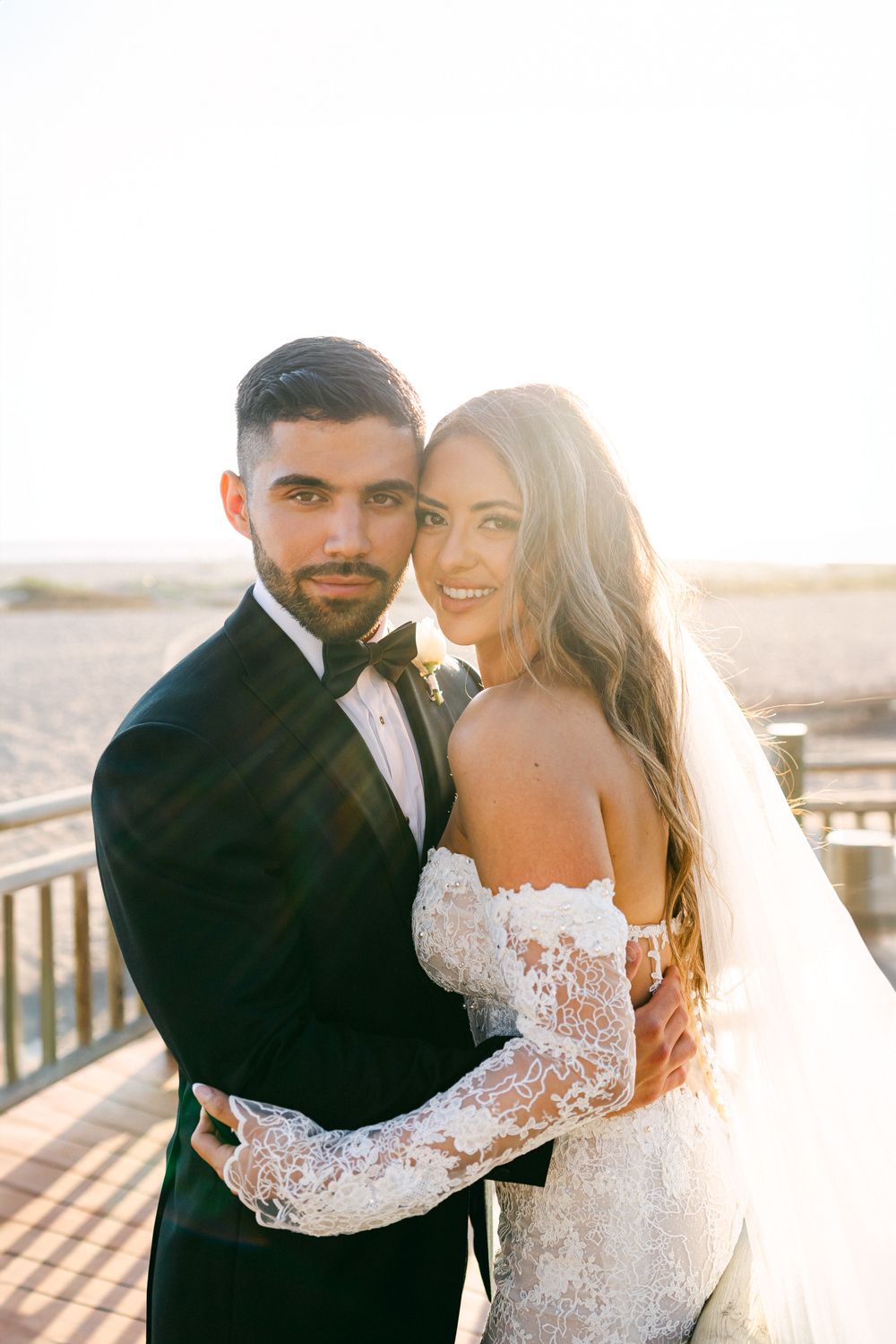 A sun-drenched wedding portrait features a couple in elegant attire on a wooden deck.