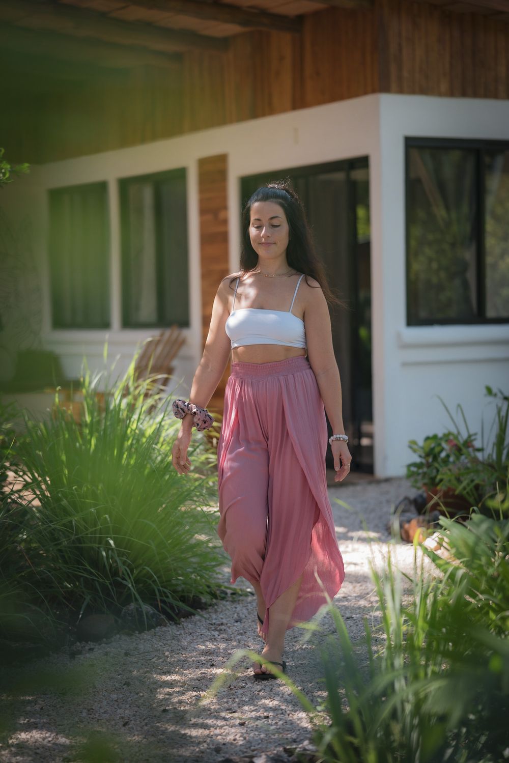 Person wearing a white crop top and flowy pink skirt walks along a garden path on a sunny day.