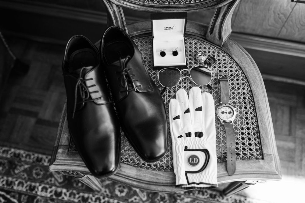 Black and white detail shot of groom's shoes and accessories on patterned surface.